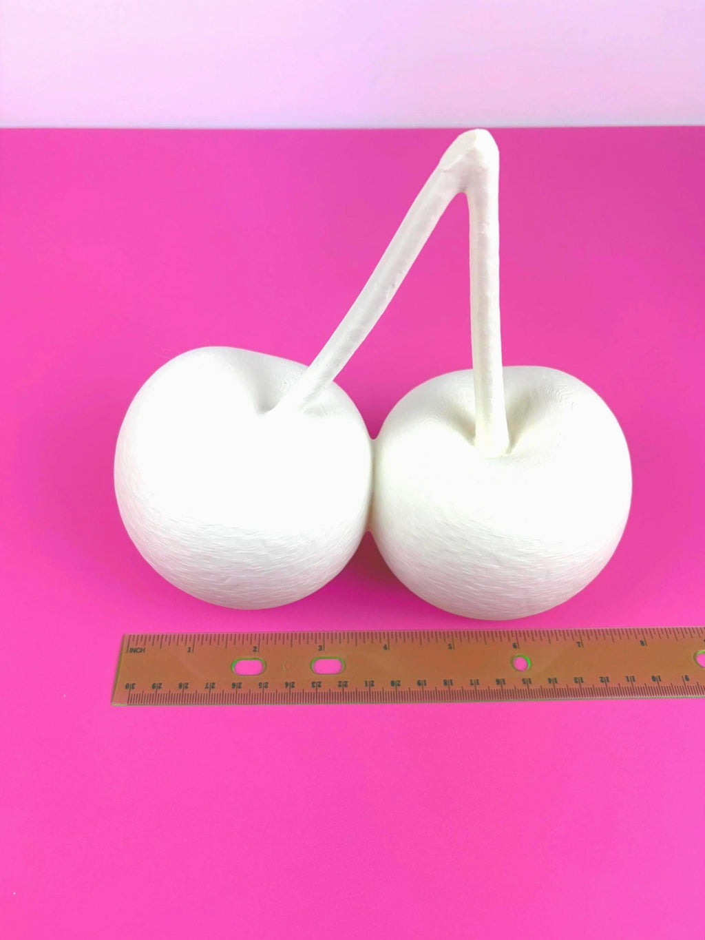 May include: cherry 3D model connected by a curved stem, resembling cherries. The objects are set against a vibrant pink background. A ruler is placed near the objects for scale.
