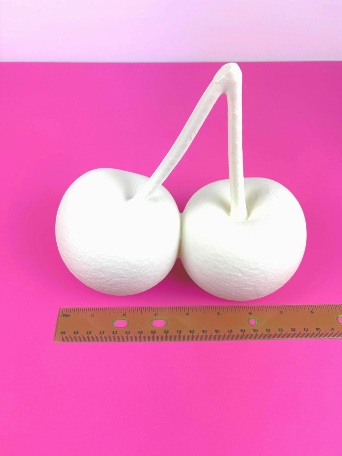 May include: cherry 3D model connected by a curved stem, resembling cherries. The objects are set against a vibrant pink background. A ruler is placed near the objects for scale.